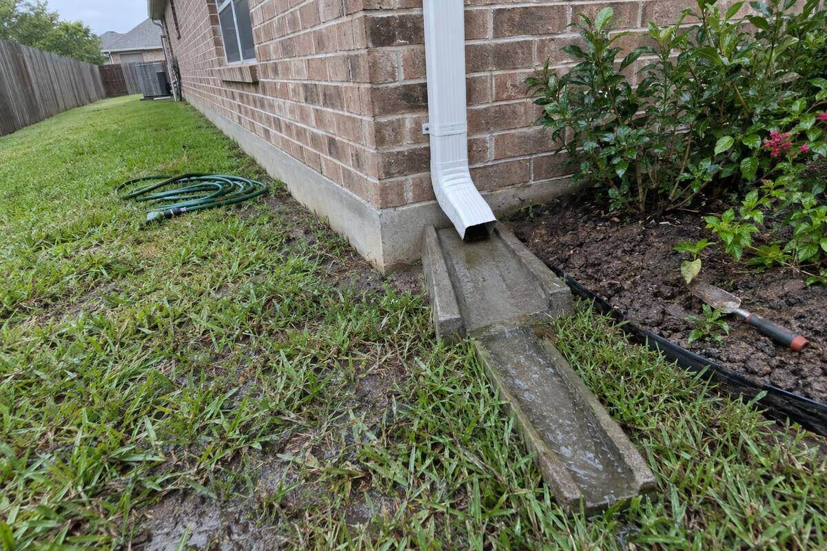 White seamless gutter and downspout at the corner of a brick house, draining onto a concrete splash block beside the slab