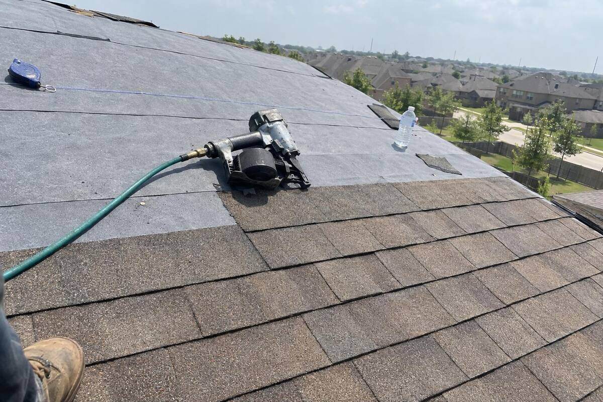 Rows of laminated architectural shingles partway up a roof slope with a roofing nailer lying on them